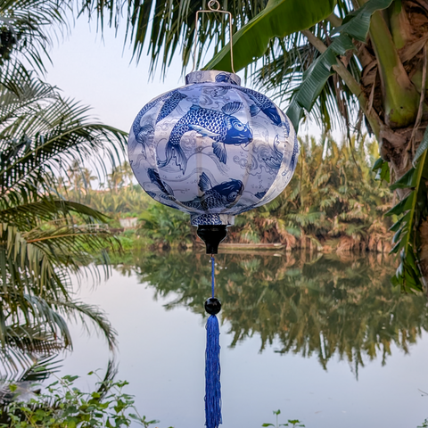 Round lantern hanging in front on a river and boat amongst palm leaves with a print of Blue Koi fish a pattern of intricately detailed koi fish swimming amidst silver swirling water currents.