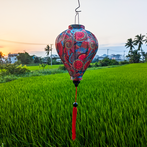 Balloon shaped Vietnamese lantern with red tassel in a rice field at sunset with red, blue, orange, green and yellow paisley print.