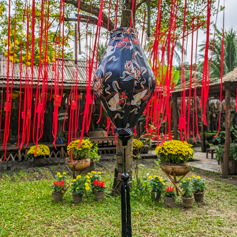 Tube shaped Vietnamese Hoi An Lantern in a garden setting with red ribbons and pretty yellow flowers.