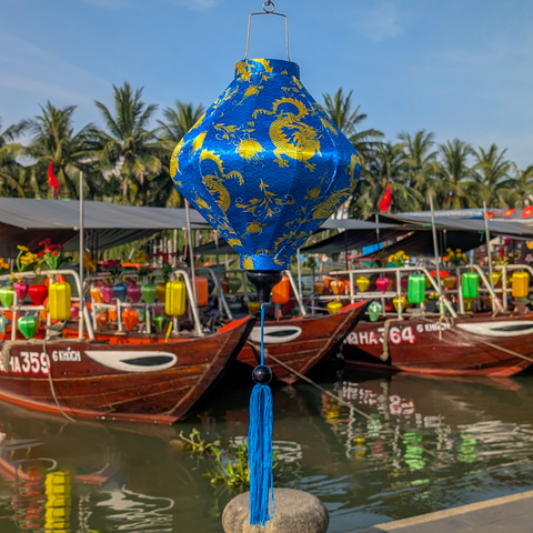 Decorative blue lantern with yellow patterns hanging above boats on a waterway with palm trees in the background.