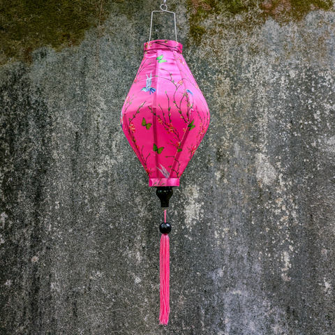 Tube shaped lantern with pink tassel and print of hummingbirds mid-flight hunting nectar from orange flowers on pink background hanging in front of a grey moss covered wall.