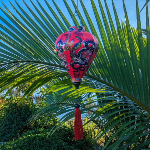 Balloon shaped lantern with red tassel adorned with intricate prints of dragons, peacocks, and butterflies against a striking red background set in front of a river with large palm leaves.