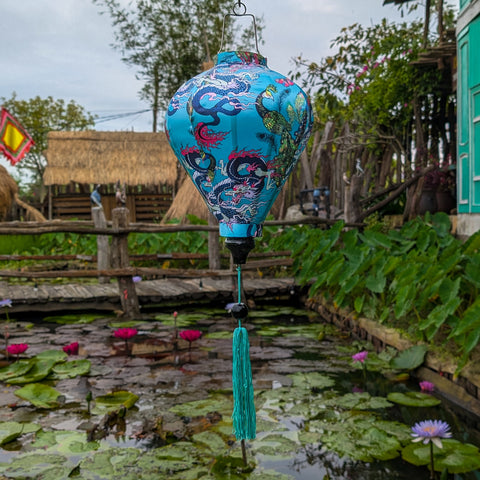 A striking blue Hoi An silk lantern adorned with intricate dragon and peacock motifs in vivid shades of pink, purple, and turquoise. The detailed artwork showcases traditional Vietnamese craftsmanship, complimented by a matching teal tassel. Hanging gracefully over a tranquil lotus pond, the lantern creates a serene and cultural ambiance.