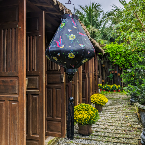 Teardrop shaped Vietnamese Hoi An lantern with a print of flying swallows with striking red and blue wings set against a bold black backdrop adorned with diamonds and yellow daisy flowers in a garden with a row of brown doors and beautiful flowers in the background. Perfect decor for the garden!