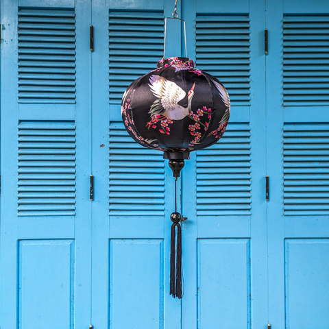 Round shaped Vietnamese Hoi An lantern featuring red-headed white cranes amongst pink cherry blossoms against a black background hanging in front of blue shutters. Perfect for home decor.