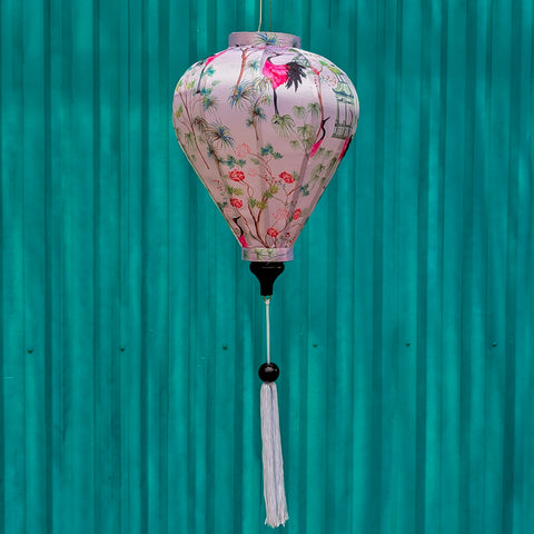 A balloon shaped lantern with white tassel hanging in front of a painted blue wall featuring pink and black cranes among lush green foliage and delicate flowers.