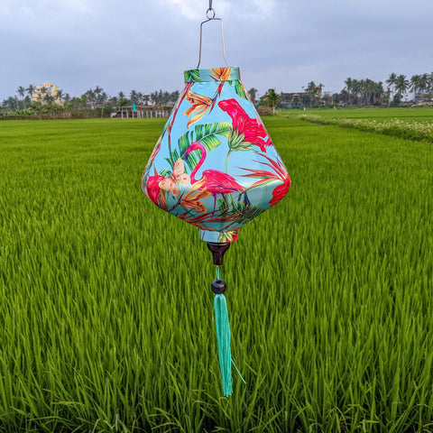 Teardrop shaped lantern with a print of pink flamingos on a light blue background hanging in a rice field.