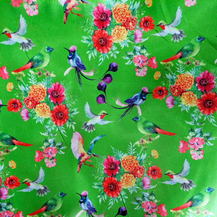 Close up of polyester silk fabric with a print of bright bunches of flowers and various feathery avian species on a bright green backdrop.