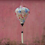 A balloon shaped lantern made with bamboo and polyester silk fabric featuring graceful cranes, vibrant chrysanthemums, and delicate dragonflies hanging in front of a pink grimey wall.