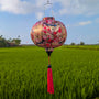 Round shaped lantern with floral and birds design hanging against a green field and blue sky.