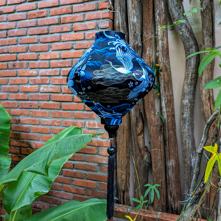 Lozenge shaped lantern with black tassel in a garden setting featuring a print of birds in graceful flight, set against a sleek black background adorned with rolling waves, floating flowers and intricate gold illustrations.