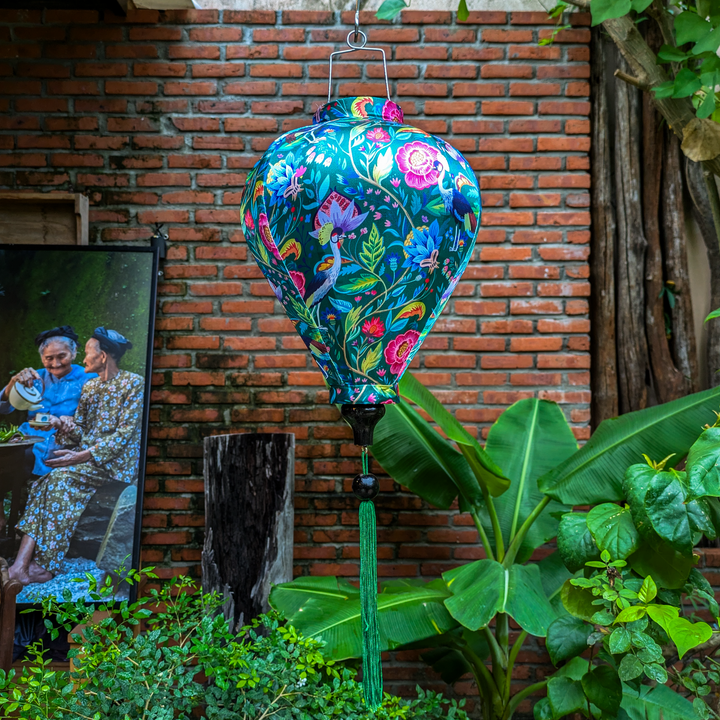 Balloon shaped lantern featuring a print of lush, tropical-inspired design on a deep green silk fabric hanging in a garden in front in front of a brick wall with plants and a picture frame depicting two Vietnamese woman drinking tea.