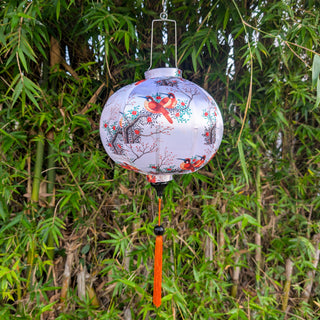 Round lantern with floral and bird design on white hanging against a green leafy background with orange tassel.