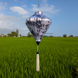 Decorative black and white diamond shaped lantern with white tassel against a green rice field and blue sky background