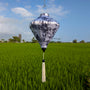 Decorative black and white diamond shaped lantern with white tassel against a green rice field and blue sky background