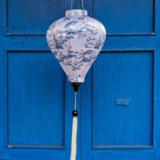 Balloon shape Vietnamese silk lantern with a illustration of bonsai trees in blue against white. Hanging in front of blue shutters in Hoi An.