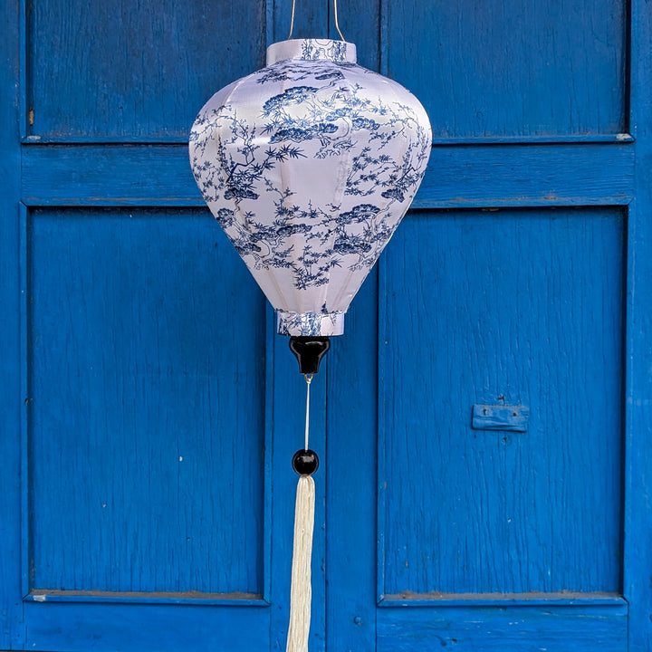 Balloon shape Vietnamese silk lantern with a illustration of bonsai trees in blue against white. Hanging in front of blue shutters in Hoi An.