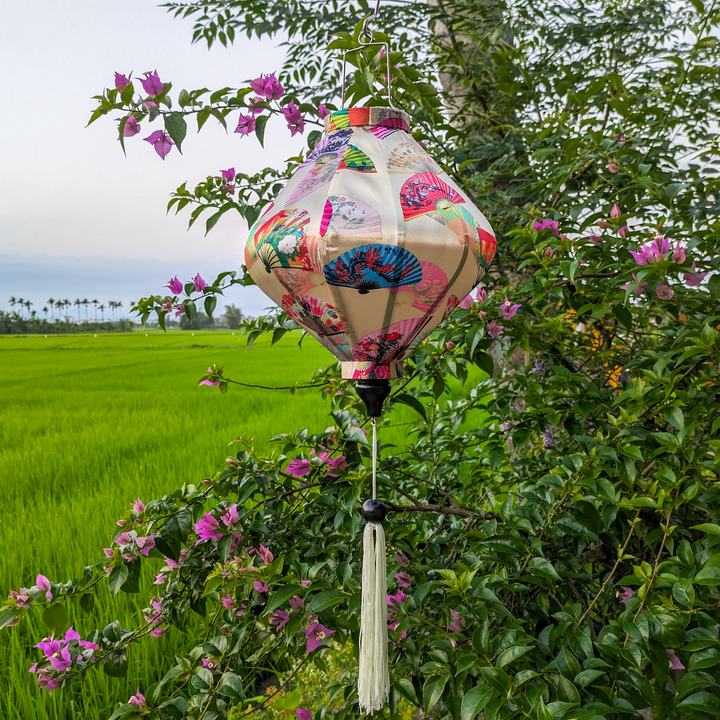 Lozenge shaped Vietnamese lantern with white tassel featuring quirky fans shaped in diverse styles and colours, seamlessly set against a captivating cream backdrop hanging in front of rice fields with palm trees.