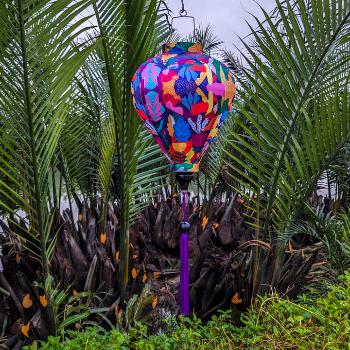 Balloon shaped lantern with purple tassel hanging outside in front of palm trees on a river.