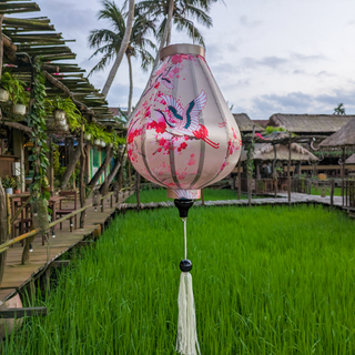 Garlic shaped Vietnamese lantern set against rice fields at Chic Chillax Cafe in Hoi An featuring majestic cranes in vivid reds, hints of teal and delicate blossoms against a calming creamy backdrop. 