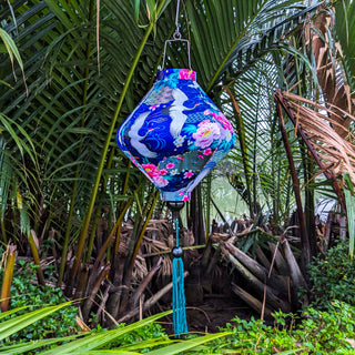 Lozenge shaped lantern with blue tassel hanging outside featuring white cranes flying amidst a dark blue background of geometric shapes, flowers, and golden clouds. 
