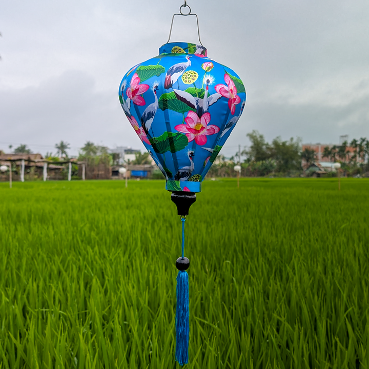 A vibrant blue Hoi An silk lantern featuring elegant white cranes, lush green lotus leaves, and striking pink lotus flowers. The intricate hand-painted design adds a touch of nature and tradition, while the matching blue tassel enhances its graceful aesthetic. Set against a backdrop of lush green rice fields, this lantern beautifully represents Vietnamese craftsmanship and cultural artistry.