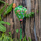 A vibrant green Hoi An silk lantern featuring elegant white cranes in flight, delicate pink and red peonies, and intricate floral details. The lantern hangs gracefully in a lush tropical setting with a rustic wooden backdrop adorned with artistic terracotta masks. A matching green tassel dangles from the base, enhancing its traditional charm.