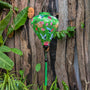 A vibrant green Hoi An silk lantern featuring elegant white cranes in flight, delicate pink and red peonies, and intricate floral details. The lantern hangs gracefully in a lush tropical setting with a rustic wooden backdrop adorned with artistic terracotta masks. A matching green tassel dangles from the base, enhancing its traditional charm.