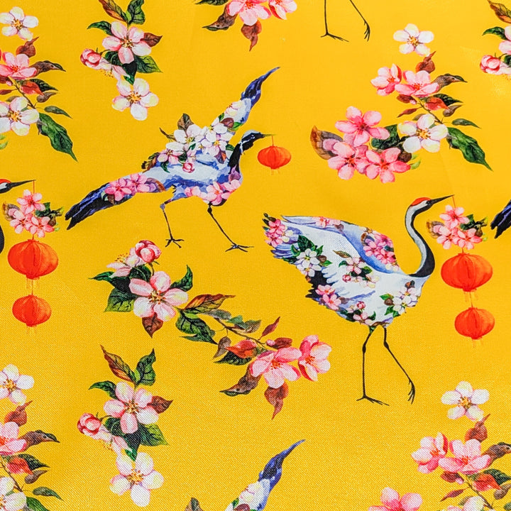Polyester silk fabric with a print of red headed cranes with flowers on their wings holding lanterns in their beaks on yellow.