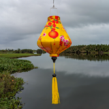 Teardrop shaped Vietnamese lantern with a yellow tassel set against a river with palm trees and green grass.