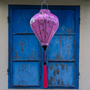 Balloon shaped Vietnamese Hoi An lantern with a pink tassel hanging in front of blue shutters.