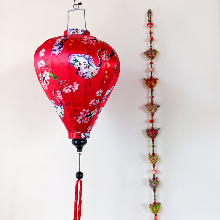 Balloon shaped lantern with red-necked birds and round lanterns, surrounded by pink and white flowers on a red background in front of a wall next to hanging decoration.