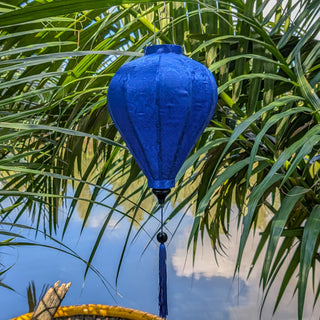 Blue jacquard silk lantern hanging among palm leaves with a blue sky and clouds reflecting in the river. 