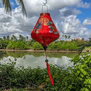 Teardrop shaped lantern with red tassel featuring a print of green and blue dragons along with golden symbols on red silk hanging outside in front of the Thu Bon River.