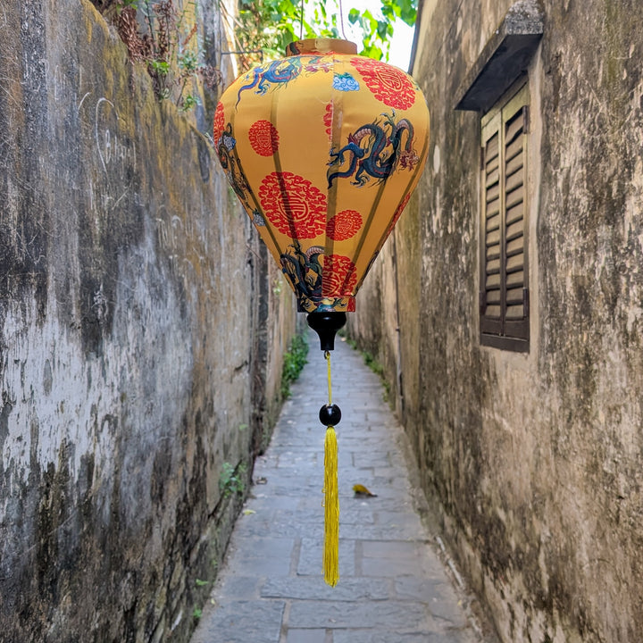 Balloon shaped lantern featuring dragons on yellow background hanging in a narrow alleyway with textured walls in Hoi An Old Town, Vietnam.