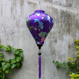 A balloon shaped lantern hanging in front of a grey wall in a garden featuring a print of koi fish swimming around bamboo reeds and pink lotus flowers on purple background.