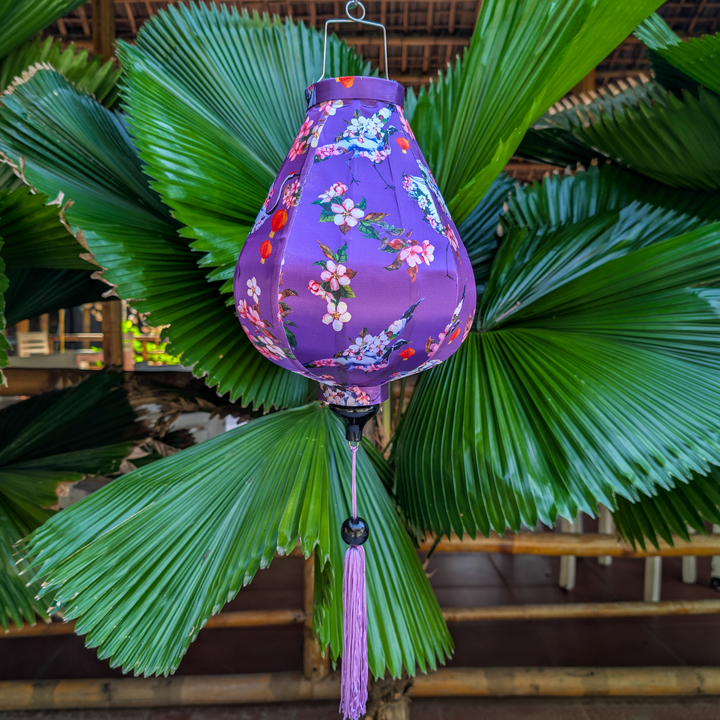 Garlic shaped purple floral lantern with birds holding lanterns in their beaks hanging among green leaves with a matching purple tassel