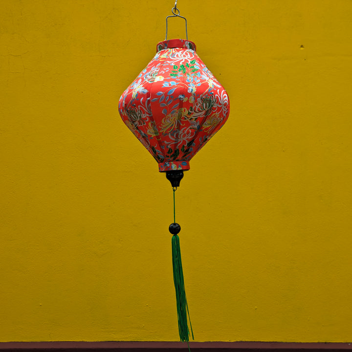 Lozenge Vietnamese lantern with delicate chrysanthemum flowers in yellow, green, brown hanging in front of a bright yellow temple wall. 