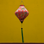 Lozenge Vietnamese lantern with delicate chrysanthemum flowers in yellow, green, brown hanging in front of a bright yellow temple wall. 