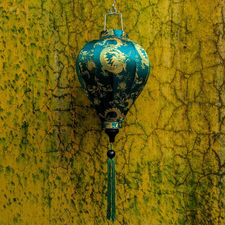 A balloon shaped lantern with a print of yellow dragons on dark green background hanging in front of a textured, cracked yellow and green wall.