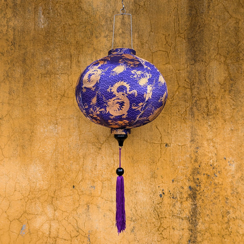 Round purple lantern with gold dragons print hanging in front of a textured old wall in Hoi An