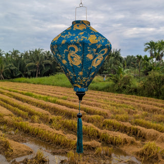 Diamond shaped Vietnamese silk lantern in a teal green with golden yellow dragons hanging over a rice field.