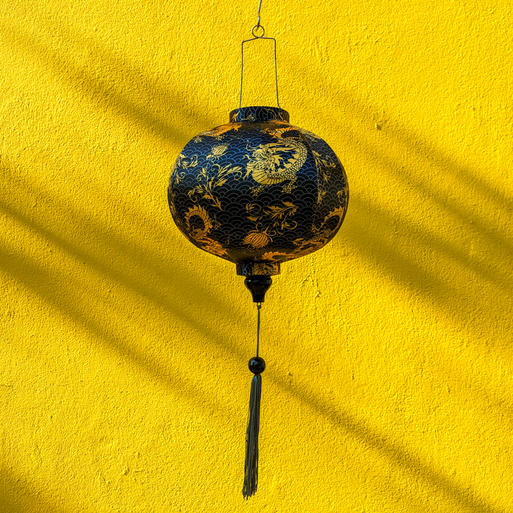 Round Vietnamese lantern with a print of golden yellow dragons and flowers set against a grey sea pattern of swirls on a rich black background set against a bright yellow wall casting beautiful shadows.