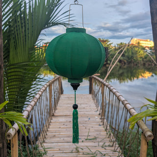A green poly linen fabric lantern with matching tassel hanging suspended over a bamboo bridge over calm water, framed by tropical plants.