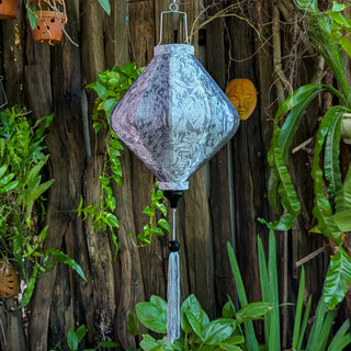 Grey jacquard silk lantern in Lozenge shape with matching grey tassel hanging in front of a wooden fence and plants