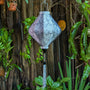 Grey jacquard silk lantern in Lozenge shape with matching grey tassel hanging in front of a wooden fence and plants