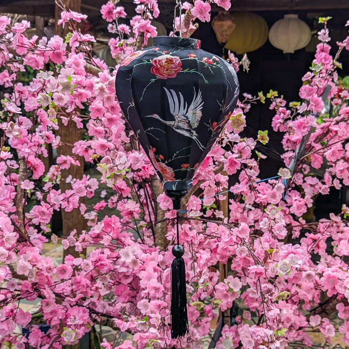 A stunning black Hoi An silk lantern featuring elegant white herons in flight, delicate pink roses, and intricate floral details. The lantern is set against a backdrop of vibrant pink cherry blossoms, creating a striking contrast. The black tassel hanging below adds to its traditional charm. This lantern embodies grace, longevity, and timeless beauty, making it a perfect decorative piece for homes, festivals, or gifts.