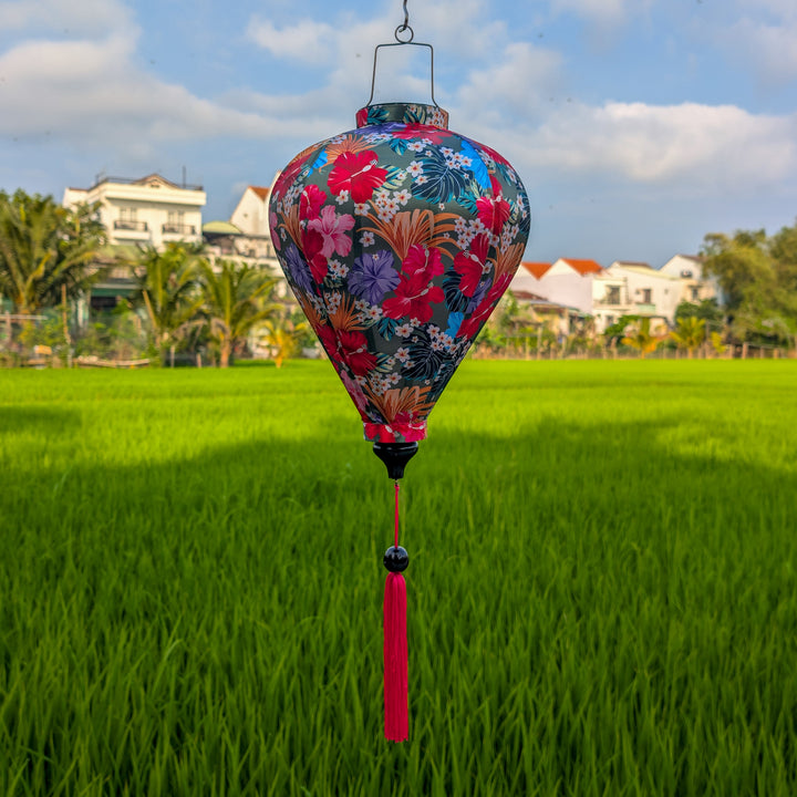 Balloon shaped handmade silk lantern with floral design and bright pink tassel hanging over a green field with houses in the background