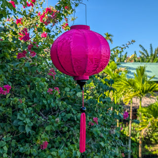Round Jacquard silk Hot Pink lantern hanging among green foliage and flowers with matching pink tassel