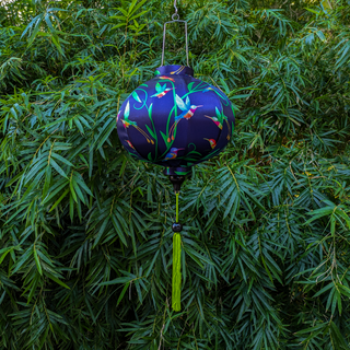 Round lantern with a print of hummingbirds and plants on dark blue silk background hanging outside in front of a large green bush.